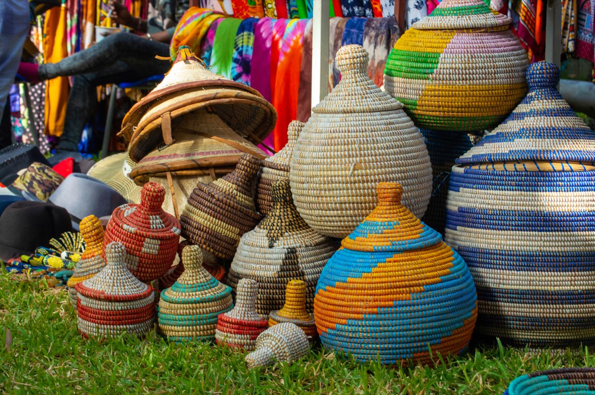 Lidded baskets of coiled grass in bands of blue, orange, red and cream, gathered on the grass at an outdoor market.
