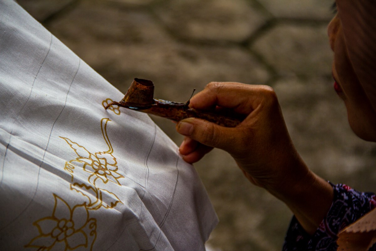 A hand guides a small copper batik tool, drawing a line of wax onto white cotton already patterned with golden flowers.