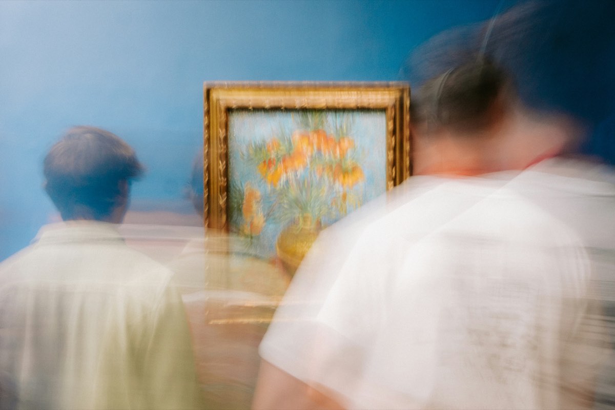 Visitors move past a gilt-framed painting of orange flowers on a blue gallery wall, their figures softened by motion blur.